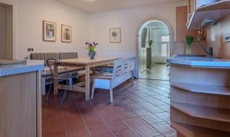 An inviting dining area with a large table, chairs, and decorative pictures on the wall. The windows let in plenty of light and the floor is designed with terracotta-colored tiles.