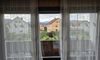 A view from a window with white curtains. Outside, colorful houses and green landscape can be seen.