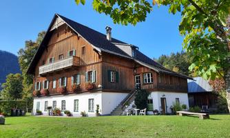 A traditional wooden house with a large garden and many windows. In the background, trees and a blue sky are visible.