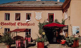 A traditional inn with the sign "Gasthof Eberhard". In front of the building, there are tables and chairs under red sun umbrellas.