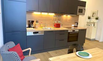 A modern kitchen with dark cabinets and a light wood countertop. In the foreground is a table with a green plate and a chair with a red cushion.