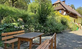 A cozy outdoor area with a wooden table and benches, surrounded by green plants and flowers. In the background, a two-story house can be seen.
