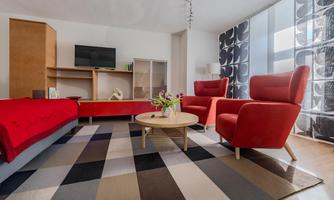 A modern living room with red armchairs and a round table. The room has large windows and a carpet in neutral colors.