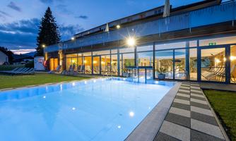 A modern outdoor pool with smooth water and an elegant terrace. In the background is a building with large windows, illuminated at dusk.