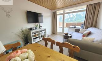 A modern living room with a cozy sofa and a television. Large windows offer a view of the mountains.