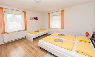 A bright bedroom with two beds and orange decoration. The floor is made of wood, and there are large windows with curtains.