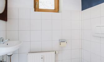 A modern bathroom with white tiles and a small window. There is a sink, a heater, and a toilet with a special toilet seat.