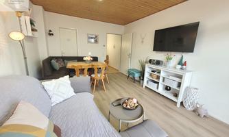 A bright and modern living room with a cozy sofa and a dining table. There is a television on the wall, and the wooden ceiling gives the room a warm atmosphere.