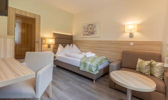 A cozy hotel room with a double bed and a seating area. It is bright and modernly furnished, with warm wood tones and soft colors.