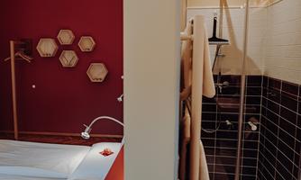 A modern bedroom with a red painted wall and hexagonal shelves. In the background, a shower cabin is visible.
