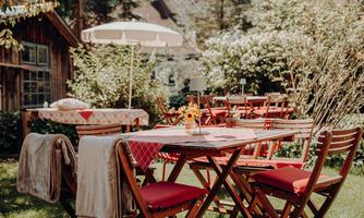 A cozy garden with wooden tables and red chairs. A sun umbrella provides shade while flowers and trees beautify the surroundings.