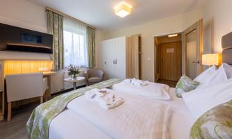 A modern hotel room with a large double bed and a cozy seating area. The room is bright and friendly with windows that let in daylight.