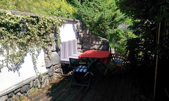 A cozy outdoor area with a wooden terrace and a red table. Surrounded by green plants and a stone wall.