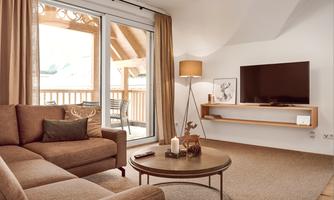 A cozy living room with a beige sofa and a round coffee table. Large windows let in plenty of light and provide access to a balcony.