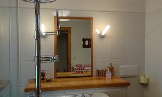 A modern bathroom with a sink, a mirror, and a shower. The walls are light tiled, and there is a wooden workspace.