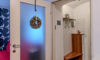 A bright hallway with a glass door and a decorative wreath. Next to the door is a wooden shelf and in the background, there are plants.