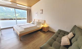 A cozy bedroom with a large bed, a sofa, and a beautiful view of the mountains. The bright room has wooden details and plenty of daylight.