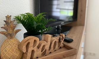 A stylish living area with a television and a wood decoration that shows "Relax". On the table, there is a pineapple and a small green plant in a pot.