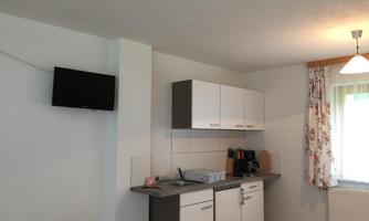 A modern kitchen with white cabinets and a small washing area. A television is mounted on the wall, and a window with curtains provides daylight.