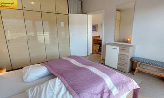 A modern bedroom with a large bed, subtle furniture, and a light color tone. The walls are light, and a large mirror adds extra brightness.