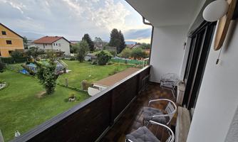 A balcony with two chairs and a view of a garden. In the background, several houses and a cloudy sky can be seen.
