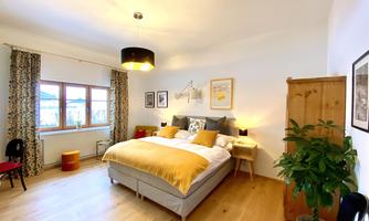An inviting bedroom with a large bed and warm colors. The room is bright and has a decorative plant accent.