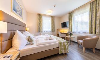 A modern hotel room with a large bed and elegant furnishings. There is a desk, a television, and large windows that provide plenty of light.