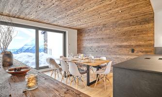 A modern dining room with wooden paneling and large windows. A table for six people stands in the center, offering a view of a winter landscape.