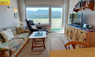 A cozy living room with a sofa and a table. Large windows offer a view of the mountains and the green landscape outside.