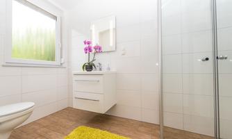 A modern bathroom with a shower and a white vanity. On the windowsill, there is a pot with orchids, and the floor is covered with a yellow rug.