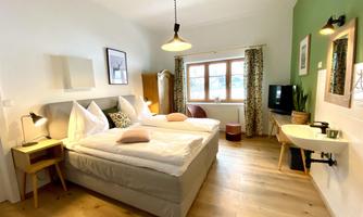A cozy room with two beds and hardwood floors. The walls are decorated in soft colors and there are large windows with curtains.