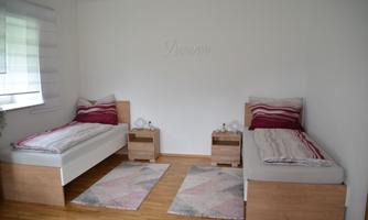 A bright room with two single beds and modern nightstands. Windows let in plenty of light and the walls are adorned with neutral colors.