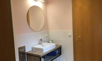 A modern bathroom with a white sink and a round mirror. The walls are painted in a soft pink, and there are wooden details on the door.