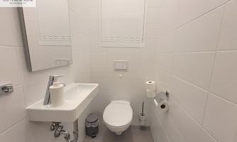 A modern bathroom with white tiles and minimalist decor. It features a sink, a toilet, and a mirror.