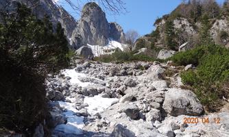 A rocky path with snow-covered spots winds through an alpine landscape. In the background, steep mountains rise under a clear blue sky.