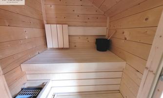 A cozy sauna with wood paneling and a lying area. On the left side, heating stones and a bucket for water are visible.