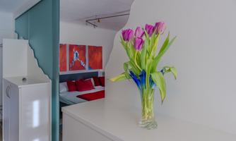 A fresh bouquet of purple tulips is in a clear vase on a white table. In the background, red wall paintings and a bedroom are visible.