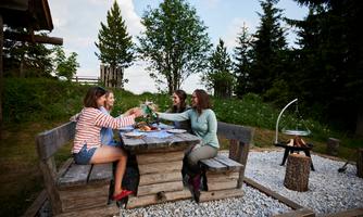 Three women are sitting at a rustic table outdoors, toasting with glasses. The table is laid with food and drinks, surrounded by a green landscape.