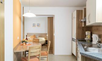 A modern kitchen with a dining area and bright wooden furniture. The room is warm and inviting, with an open access to a bedroom.