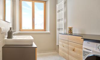 A modern bathroom with a sink, wooden cabinets, and a washing machine. Large windows let in plenty of daylight.