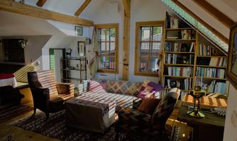 A cozy living room with wooden beams and large windows. The furnishings include various chairs, a coffee table, and bookshelves.