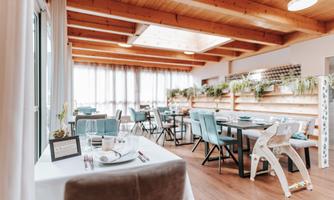A bright restaurant with a wooden ceiling and large windows. There are tables decorated with stylish chairs and plants.