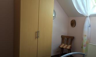 A cozy bedroom with a wooden wardrobe and a wooden chair. The window lets in plenty of light and has a white curtain.