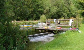 A quiet water spot with a wooden platform and green bank areas. In the background, there are trees and swimming rings visible.