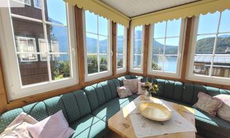 A bright conservatory with large windows and a view of a lake. A cozy seating area with cushions and a wooden table invites relaxation.