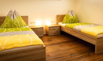 A bright room with two single beds and colorful bed covers. Next to the beds are nightstands with warm light.