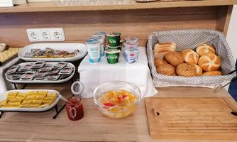 A diverse breakfast buffet with jams, yogurt, rolls, and fruit salad. The dishes are neatly arranged on a wooden shelf.