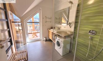 A modern bathroom with a large shower and bright tiles. In the background, there is a window with a view outside and a washing machine.