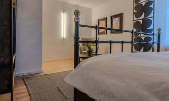A modern bedroom with a black metal bed and light walls. In the background, there are some pictures and a cozy seating area.