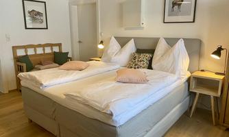 A cozy bedroom with a double bed and white bed sheets. Pictures hang on the wall and there is a small sitting area with a chair.